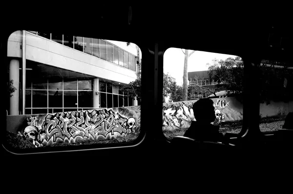 A black and white photograph showing a view from inside a Melbourne train carriage. The interior is dark, with one seated passenger visible. Through a set of windows, passing graffiti murals are visible.