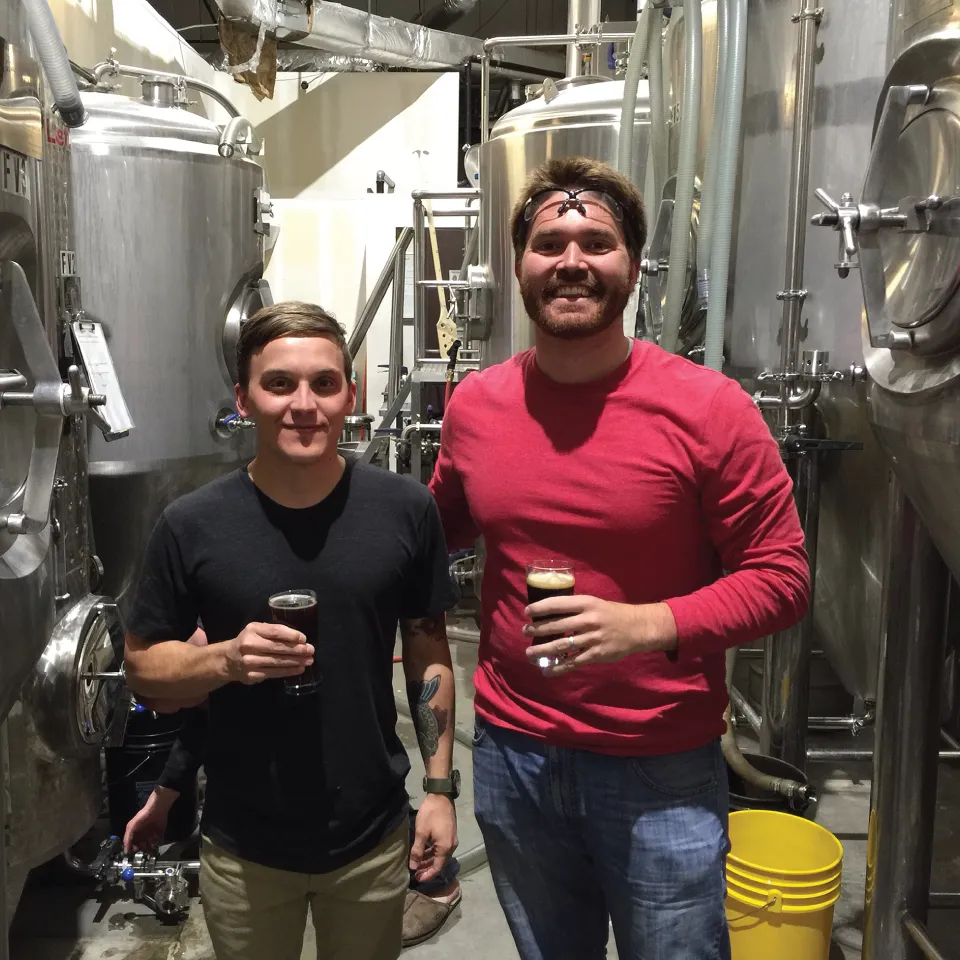 Photograph of two men with beer glasses in their hands in the processing unit of a brewing company.