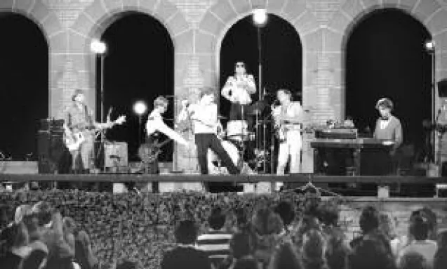 A photo of the Dishes playing in front of the stone arches at the Hart House Quandrangle. The audience is in the foreground of the photo, sitting on chairs lower than the stage. The band is on an elevated stage in front of the stone arches. The dark night sky can be seen through the arches while large spotlights shine on the band. Instruments being played by the members include a saxophone, drums, bass and electric guitar and keyboard.