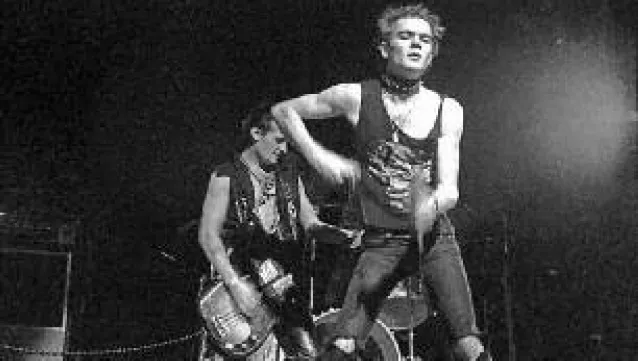 A photo of Steven Leckie with a member of Viletones behind him. Steven is wearing a muscle shirt, ripped black jeans and a large choker. The member behind him is wearing a vest without a shirt underneath and many necklaces. He is playing a fender precision bass.