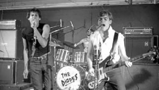 A photo of three of the members of the Diodes. Two of them are in the foreground of the photo singing into mics, one is holding a Rickenbacker bass. The drummer is behind them on a drum set that has their logo on it.