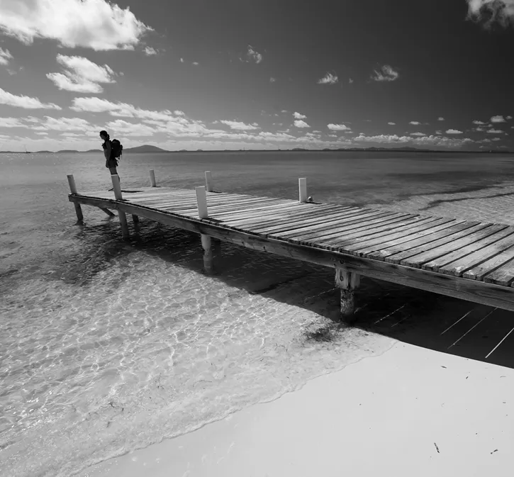 Black and white photograph of a wooden pier on a seaside beach with a young woman looking at the sea. 