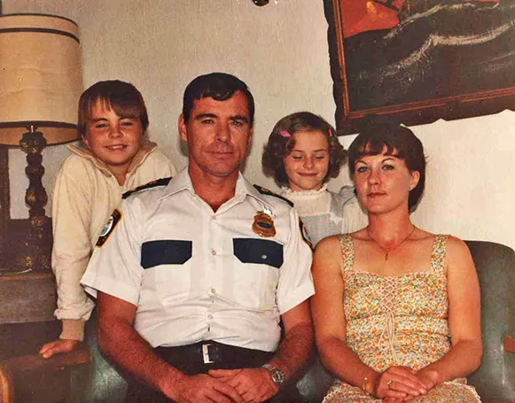 A snapshot of a family with light skin tone in their living room in approximately 1980: Chris Newton (approximately 10 years old), father Dan in his policeman’s uniform, sister Peggy (approximately seven years old), and mother Patty.