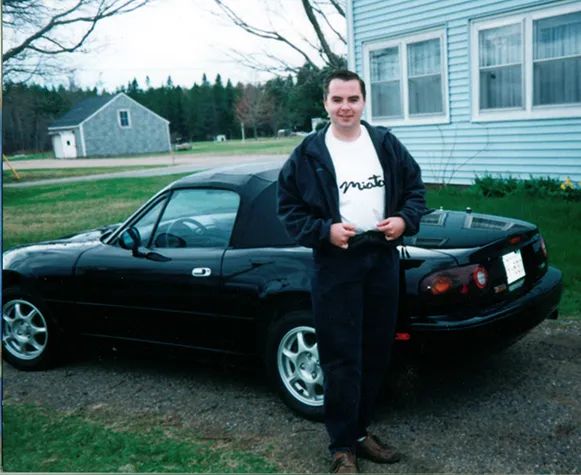 Young Chris Newton wearing casual clothes, stands beside his new, black convertible Miata sports car in the driveway of the family home. 