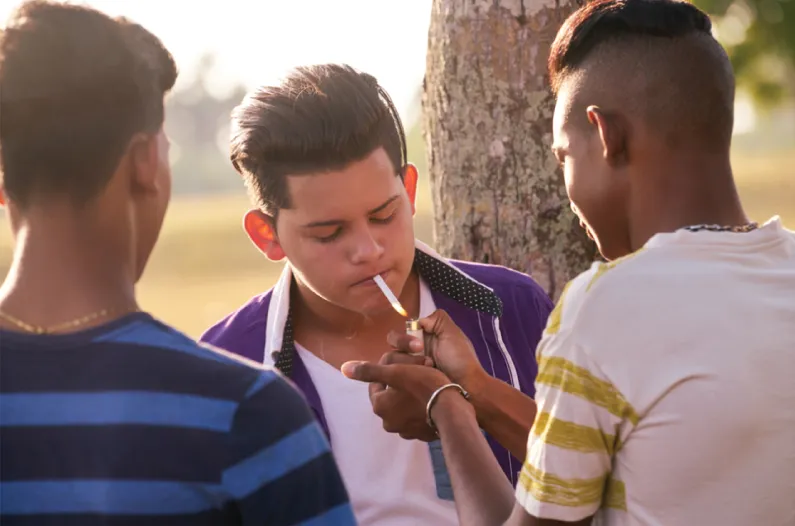 Photo depicts a three boys, among one lighting cigarette to another one.