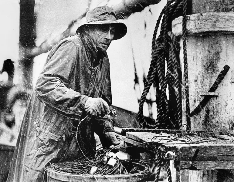Light-skinned man stands on a schooner and wears oilskins with a hat as it snows; he pulls heavy wire out of a barrel next to some rope