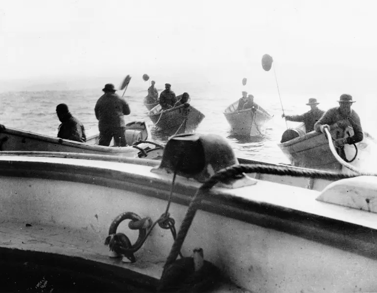 Four dories on the water that are tethered to the railing of the main vessel; each boat contains two men and one of them holds a long pole with a paddle on the end of it