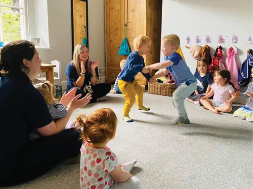 Two educators and 6 six nursery children sat in circle with two boys in centre holding hands and dancing.