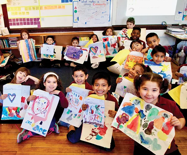 A classroom full of smiling, seated kindergarten boys and girls who are holding up their drawings and paintings.