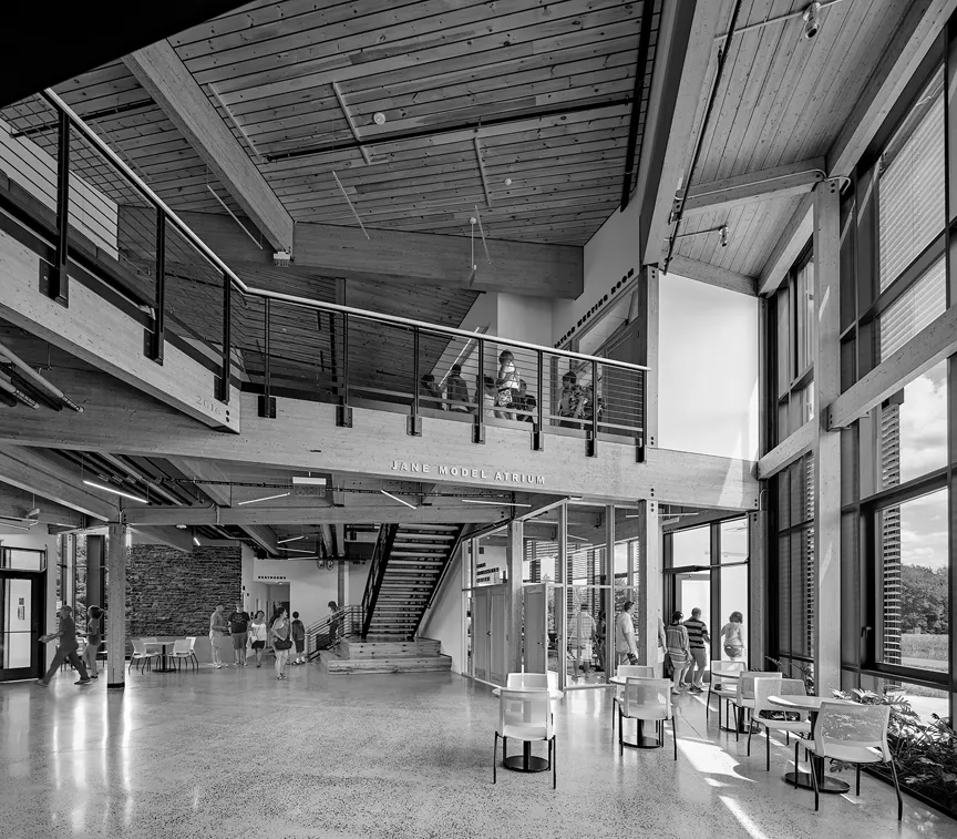 Photo of double-height interior space with a wood ceiling and exposed wood beams, people in the far right near an open stair leading to a mezzanine level with a black railing that spans from left to right, supported by timber beams and column with exposed black steel connections and column base. Underneath it an entry vestibule, on the same plane as a tall window wall on the right, with tables and chairs.