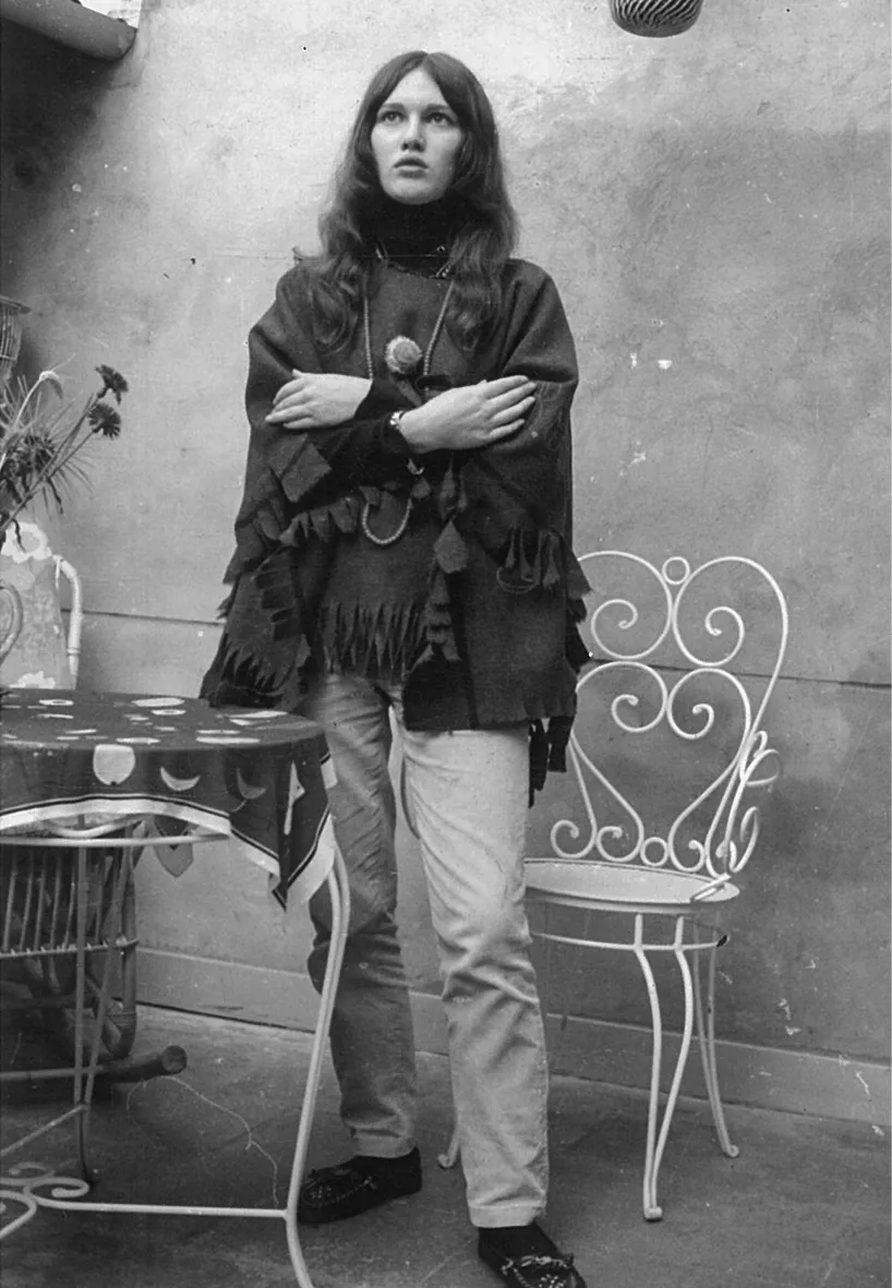 A young Diane in black-and-white, dressed in a flowing bohemian shirt, standing in a courtyard beside a table and chair 