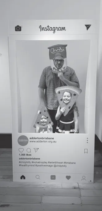 A father and his two children pose for a photo in an Instagram photo booth. The children hold crocheted watermelon artworks over their faces and the father a holds a crochet tin titled Chili Philli over his face.