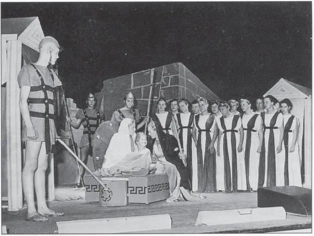 The picture is a photo of a school performance of Euripides’s play Trojan Women in 1955. On display is a stage with eighteen young actors in ancient Greek robes and soldier costumes. The set consists of two unassuming Greek temples.