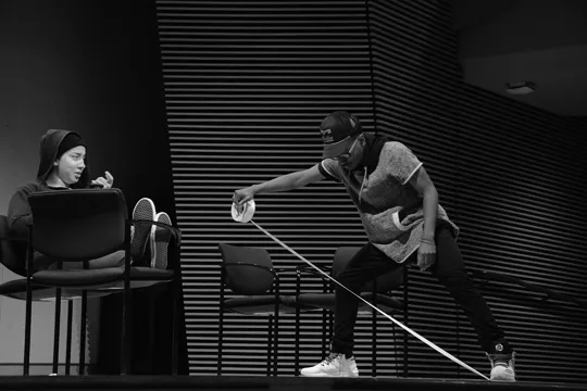 One student lays down on two chairs while another student pulls a long strip of tape across the room.