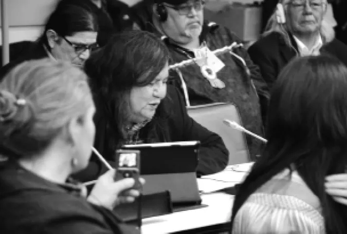 Black and white photograph of a seated Arthur Manuel speaking into a microphone at the United Nations. Manuel is surrounded by five other people. One person is recording the statement with a handheld audio device.