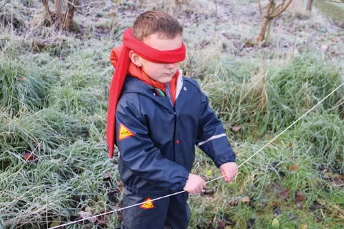 A child wearing blindfold, holding onto piece of string tied to two trees.