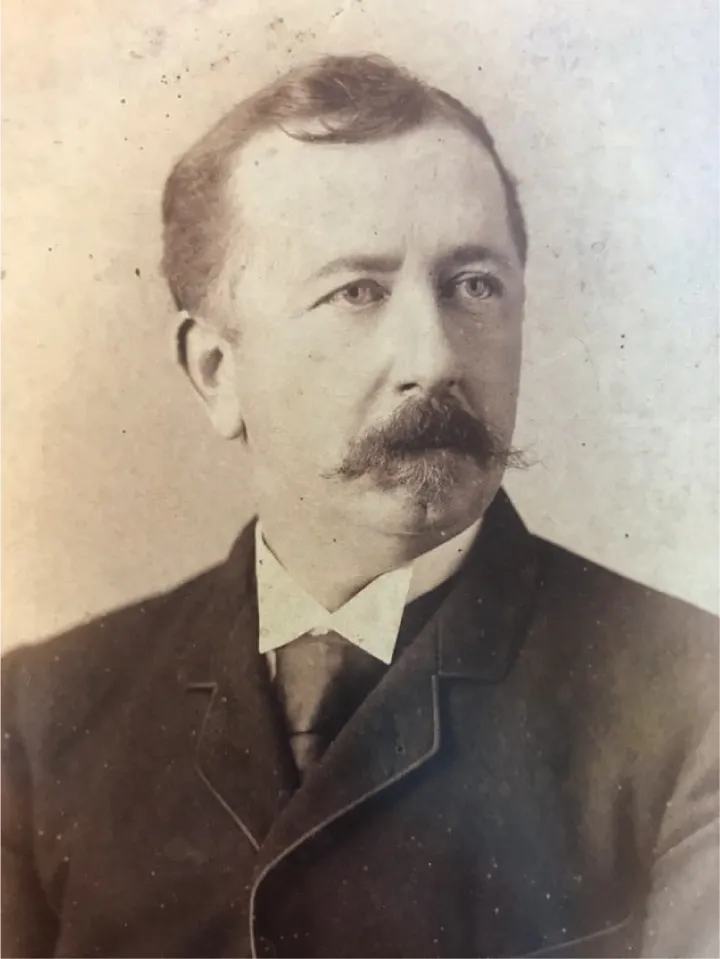 Daniel Bruton is a light-skinned man with a mustache. He is wearing a white-collared shirt and black tie with a black coat on top.