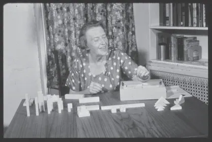 Margaret Lowenfeld is smiling sitting at a table. She is wearing a patterned dress. Curtains are behind her with a bookcase to her right-hand side. There is a heating vent underneath these blocks. Blocks of various shapes (long pieces, short pieces, cylinders and tree shapes) are on the table. There is also a wooden box with wooden shapes stacked neatly within.