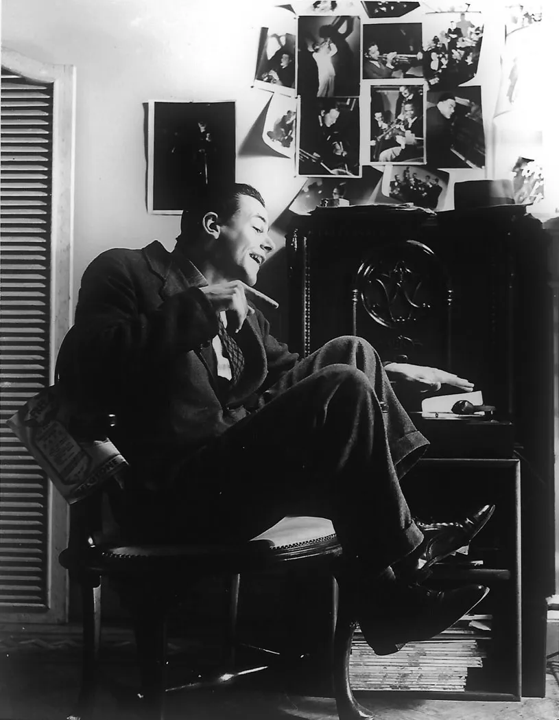 A black and white photograph showing a man in a suit listening to a record. He is sitting on a chair with his feet lifted from the ground and his finger pointing towards the record. There are numerous photographs on the wall of jazz musicians.