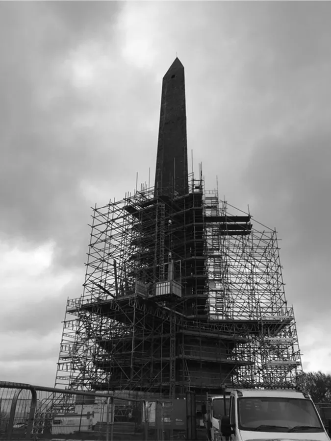 A colour photograph of Wellington Monument during its 2019 renovations, taken from the ground, close to the base, looking upwards to the pinnacle. A white works van sits in the foreground of the image with a site hut visible to the left. The three-sided obelisk that forms the monument rises through partly completed and still open scaffolding, which has reached approximately halfway up the 53m-tall structure.
