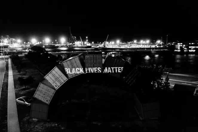 A sculpture of shipping containers arranged in an arch in a seaport context. Across the arch are projected the words: “Black lives matter.”
