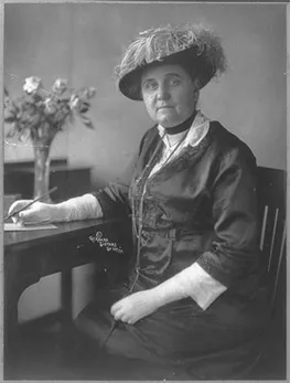 Mary Richmond posing for a picture while sitting at a table. She is holding a pen above a piece of paper on the table while looking at the camera. A vase of roses is in the background.