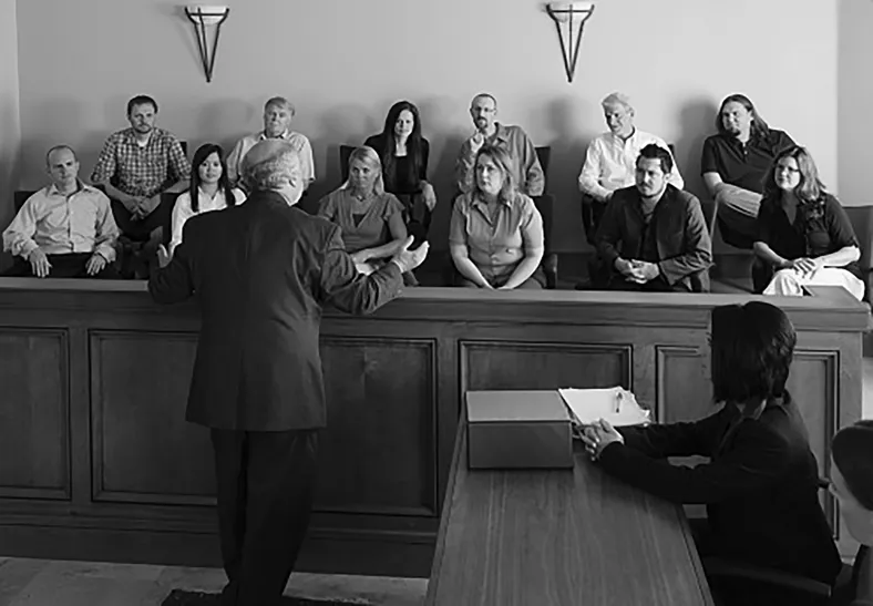 A balding man faces a jury of 12 people. The man is gesturing to the jury as he is speaking. A woman is seated to the right of him, listening to the lawyer.