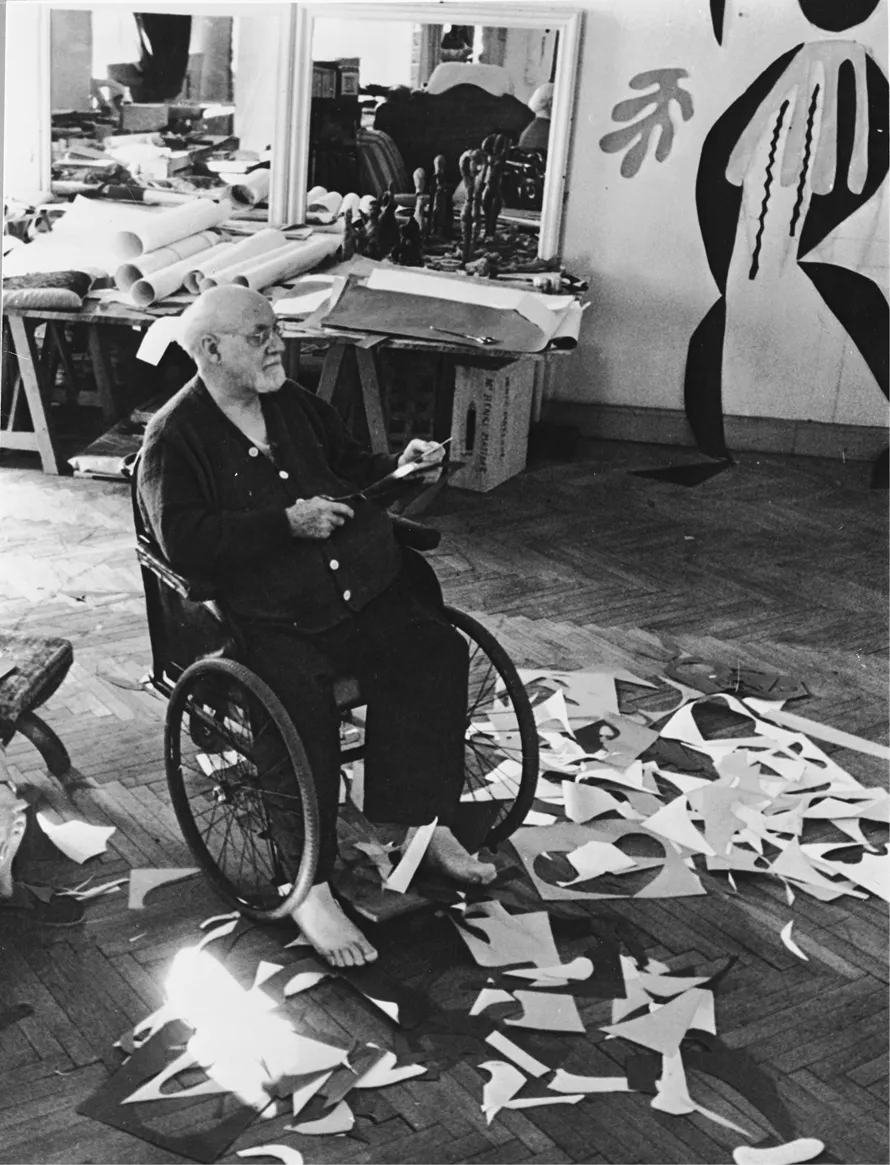 A photograph of artist Henri Matisse in a wheelchair in his studio working on cut-outs.