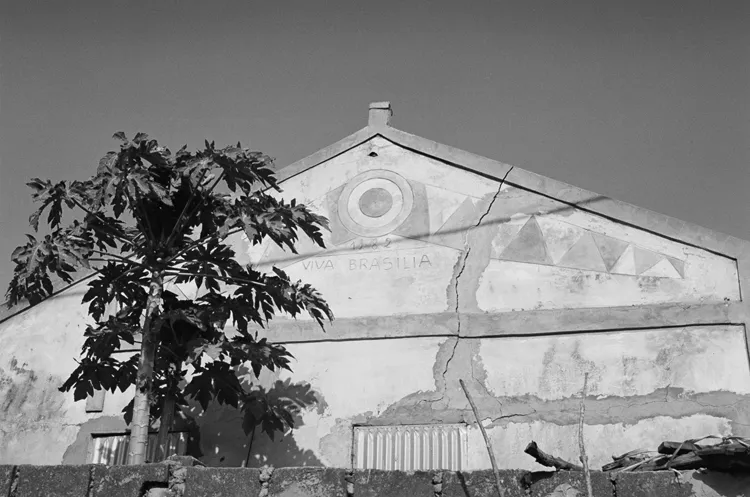 The gable of a Brazilian-style house painted yellow in a light blue frame and decorated with geometric motifs. There is an inscription saying “Viva Brasilia” with the date 1982 and a large crack runs to the right of the gable, and two openings covered in corrugated metal can be just seen above the outer wall.