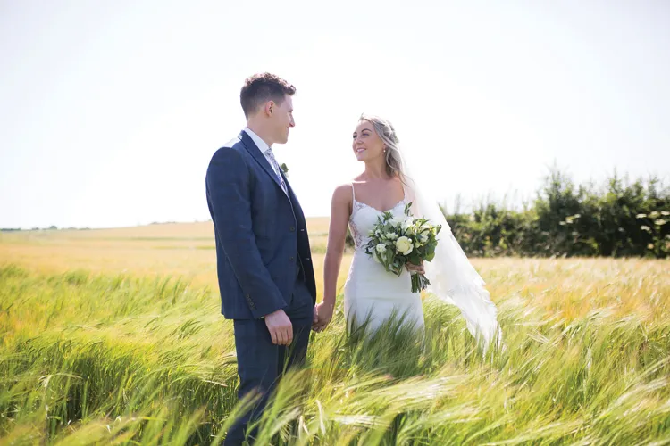 A photograph shows a bride and groom standing in a cornfield holding hands and gazing at each other. The bride holds a bouquet in her hands.