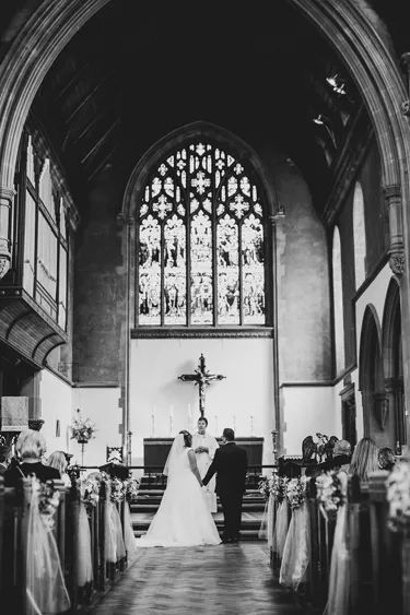 A photograph shows a wedding ceremony in a church. The bride and groom holding hands stand in front of the priest at the altar. The guests are seated in the aisles.