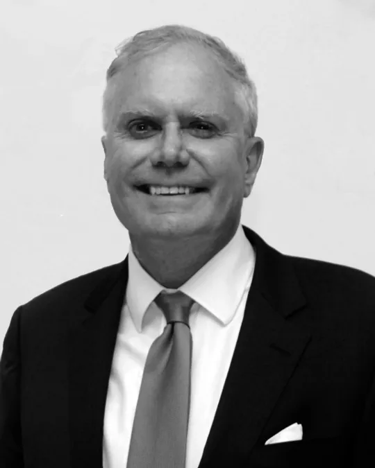 An older man with light skin tone and light grey hair smiling at the camera. He is seen from the chest up wearing a dark blazer and a white shirt with a solid-coloured tie. A white pocket square protrudes from the chest pocket of his blazer.