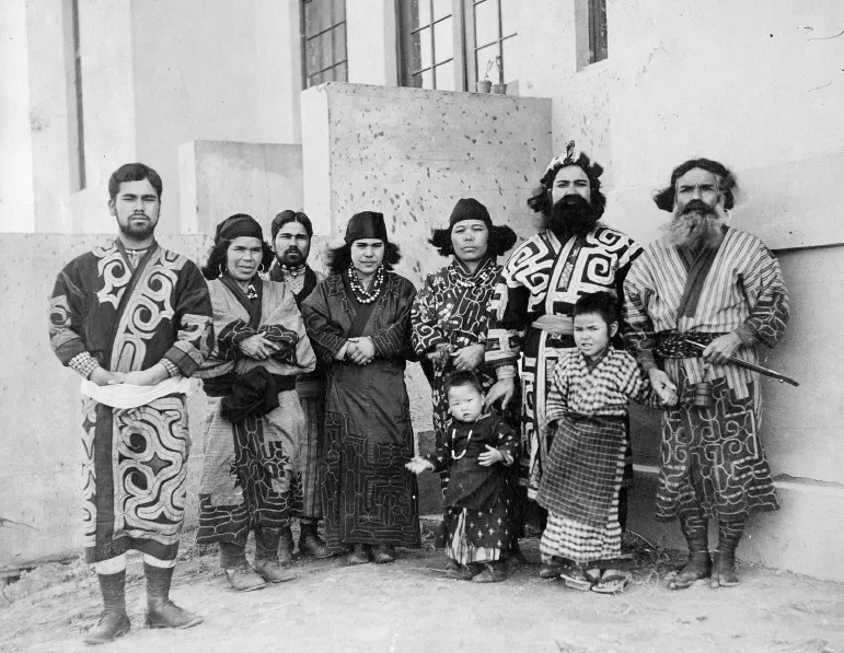  A black-and-white photo shows a group of nine Ainu people including four men, three women, and two children posing together in their traditional attire.