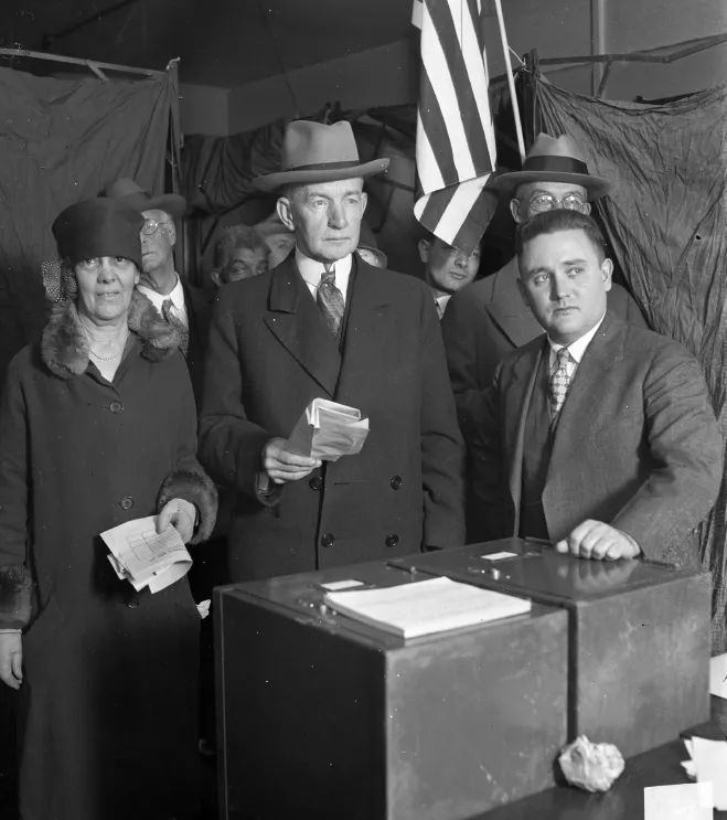 A black-and-white photo shows former US Vice President Charles Dawes and his wife posing as they are about to cast their votes. An official stands next to him, and a bunch of other dignitaries stands behind them.