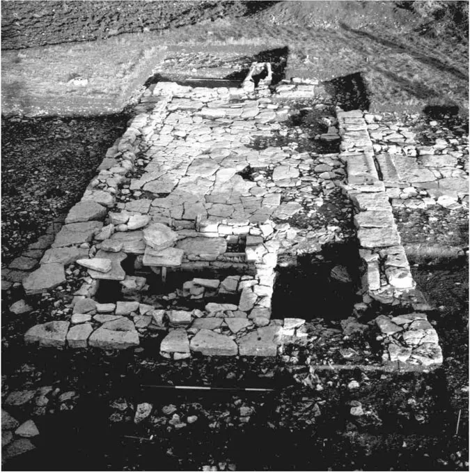 Photograph of the foundations of an excavated Romano-British stone-built barn at Worth Matravers, Dorset. At one end is a grain-dryer; hot air from the stoke-pit was drawn under the partly surviving stone-flagged floor, on to which grain would have been piled to ‘parch’ it for easier milling, or to stop it germinating if it was wanted for ale. Production of cereals in an area of thin soils, so close to the sea that salt-laden winds would have affected their growth, reflects imperial demand for British grain. The dryer was filled in and floored over; either the barn was still roofed, or it was a hard-standing for animals. After its demolition, a grave was cut through a wall, undated but probably of the same sixth- to seventh-century date as a small cemetery excavated nearby; a stone slab structure annexed to the middle of the barn’s wall may have been a cist grave, from which all trace of a skeleton had gone. The site therefore demonstrates the pressures placed on farmers by Rome, reduction in demand at the end of the Roman period, and new early medieval uses: the Decline and Fall of the Roman Empire in a Dorset field (Graham et al. 2002, 19-25; Ladle 2018, 78-78-93).