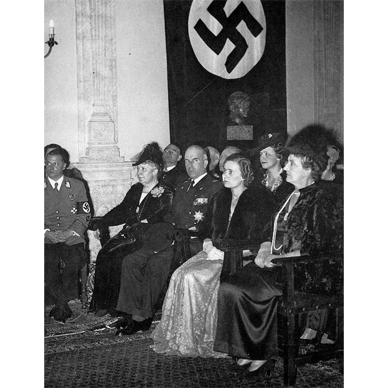 La Regina Elena (prima a destra nella foto) presenzia una cerimonia nell’Istituto Germanico di Roma. A fianco della Regina, tra due signore non identificate, il Governatore della città, principe Gian Giacomo Borghese. Sullo sfondo un gonfalone con la svastica nazista.