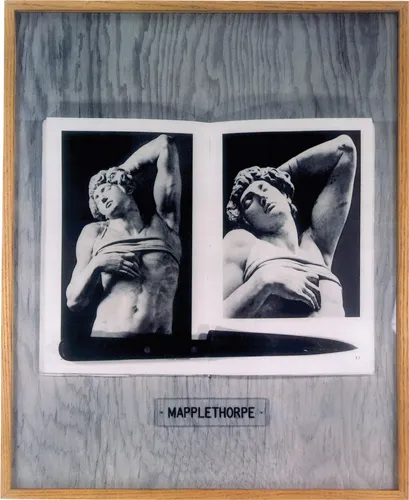 A framed photograph of an opened book on a wooden background, which is propped open by a knife. The two pages show photographs of a sculpture of a man in robes. Underneath the book is a nameplate that spells out “Mapplethorpe” in all cabs.