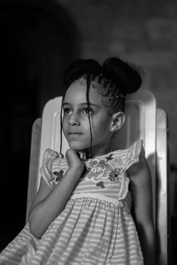 A young girl sits on a chair with her right hand on her chin. She seems to be thinking as she looks up.