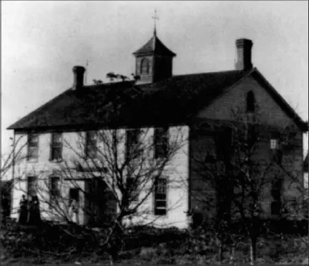 Photograph of a church at old times, it has a cross on top of the building. There are people standing out of the entrance and talking. The trees around this area have dried up.