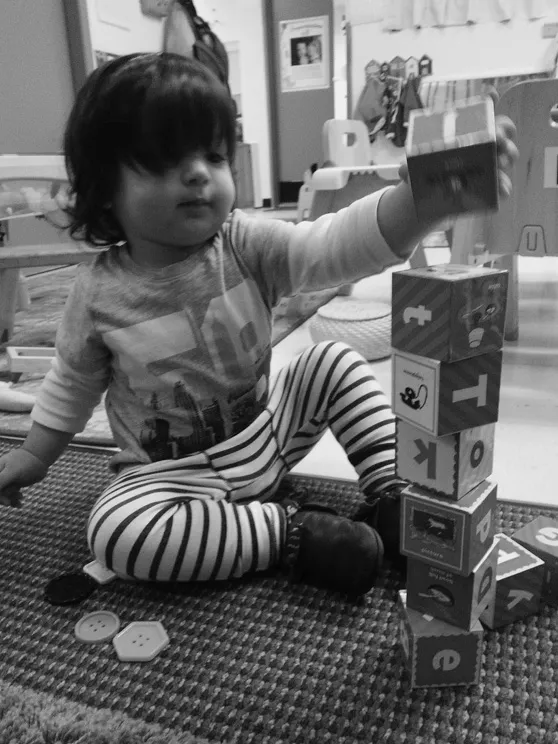 A toddler sits on the floor building a block tower as tall as she is.