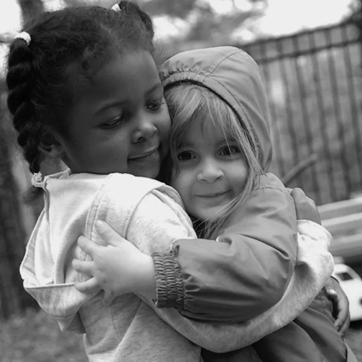 Two young girls, one Black and one White, hug on a playground while smiling.