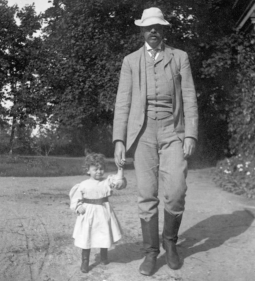 Louise as a toddler, in a puffy-sleeved, belted dress and high, sturdy boots, walking down a path with her father, a tall, thin man with droopy mustache, wearing a floppy straw hat, riding boots, and creased pale  linen suit with vest and cravat.