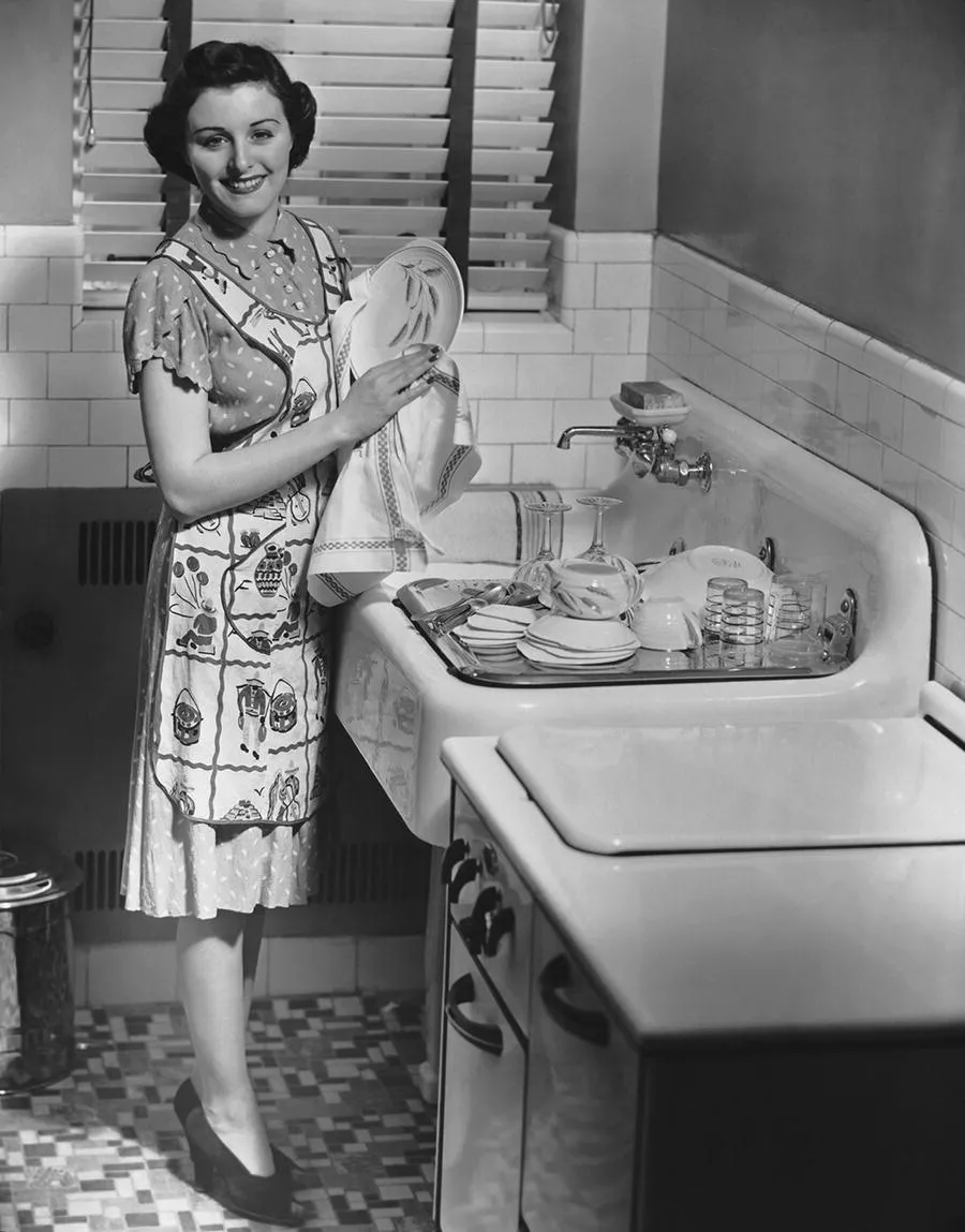 Photograph of a well-dressed woman with an apron smiling to the camera as she wipes the dishes, depicting the ideal 1950s housewife.