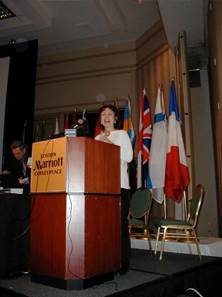 Photo of Sandra Leiblum in front of a microphone mounted on the lectern. At the background are flags of different countries beside 2 chairs.