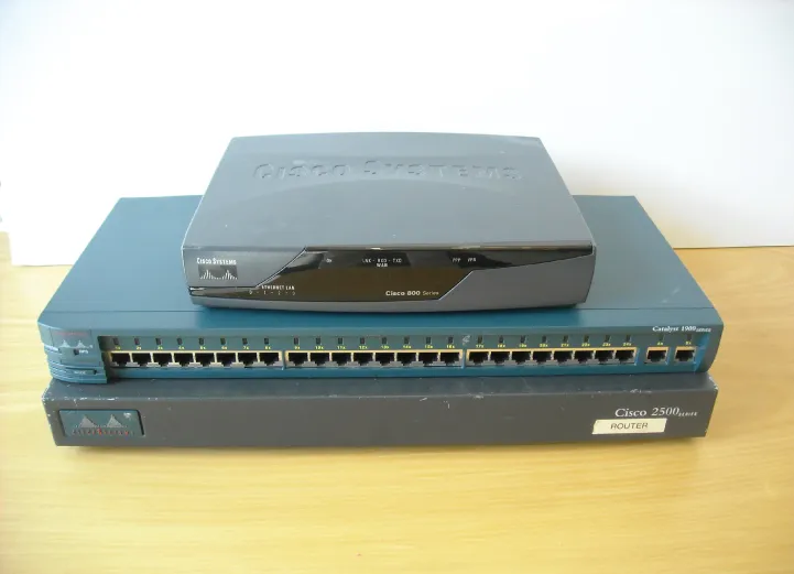 A photo of two Cisco routers and a switch in a stack: Cisco 800 series router (top), Cisco 1900 series switch (middle), and Cisco 2500 series router (bottom).